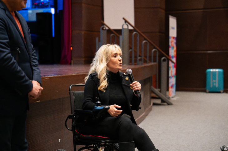 Bir salonda, sahnenin önünde tekerlekli sandalyede oturan bir kadın mikrofonla konuşma yapıyor. Üzerinde siyah bir kıyafet var, yüzü ciddi ama kararlı bir ifade taşıyor. Yanında ayakta duran biri de kısmen kadrajda. Arka planda sahne basamakları ve salonun duvarları görülüyor; ortam resmî bir etkinlik havasında.