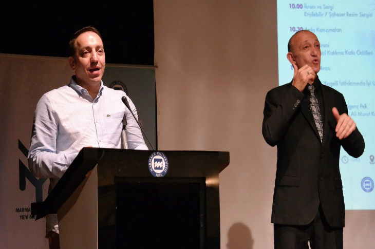 Bir sahnede, kürsüde konuşma yapan bir erkek ve yanında işaret diliyle çeviri yapan başka bir erkek görülüyor. Arkada program akışının yazılı olduğu bir sunum ekranı yer alıyor. Ortam resmi bir etkinlik havasında.