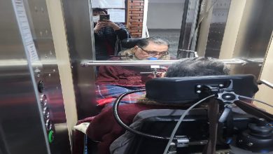 Görselde, tekerlekli sandalyede bulunan bir kişi asansör kabini içinde görülüyor. Dar alanda aynaya yansıyan asansör içi, kontrol paneli ve kabin içindeki ekipmanlar dikkat çekiyor. Sahne, asansör kullanımında erişilebilirlik ve alan kısıtını yansıtıyor.