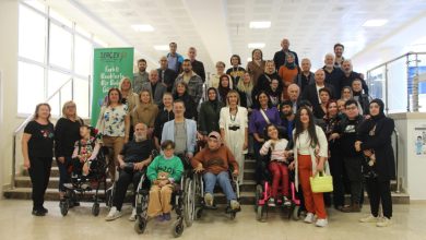 Merdivenlerde ve önünde toplanmış, aralarında tekerlekli sandalye kullanan bireylerin de olduğu kalabalık bir grup, aydınlık bir iç mekânda kameraya poz veriyor; arkada SERÇEV’e ait bir afiş görülüyor.