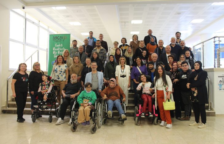 Merdivenlerde ve önünde toplanmış, aralarında tekerlekli sandalye kullanan bireylerin de olduğu kalabalık bir grup, aydınlık bir iç mekânda kameraya poz veriyor; arkada SERÇEV’e ait bir afiş görülüyor.