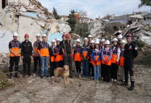 Fotoğrafta, yıkıntılarla çevrili bir alanda afet gönüllülerinden oluşan geniş bir grup yan yana dizilmiş halde poz veriyor. Çoğu turuncu yelek ve beyaz baret takıyor; aralarında arama-kurtarma kıyafeti giyen görevliler de var. Ön tarafta eğitimli bir arama köpeği dikkatle duruyor. Arka planda ağır hasarlı binaların enkazı görülüyor; sahne genel olarak bir tatbikat ya da eğitim çalışmasının yapıldığını yansıtıyor.