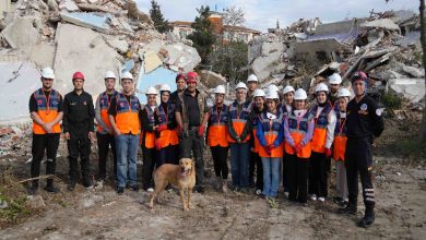 Fotoğrafta, yıkıntılarla çevrili bir alanda afet gönüllülerinden oluşan geniş bir grup yan yana dizilmiş halde poz veriyor. Çoğu turuncu yelek ve beyaz baret takıyor; aralarında arama-kurtarma kıyafeti giyen görevliler de var. Ön tarafta eğitimli bir arama köpeği dikkatle duruyor. Arka planda ağır hasarlı binaların enkazı görülüyor; sahne genel olarak bir tatbikat ya da eğitim çalışmasının yapıldığını yansıtıyor.