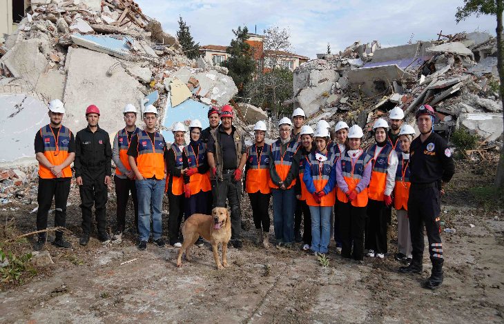 Fotoğrafta, yıkıntılarla çevrili bir alanda afet gönüllülerinden oluşan geniş bir grup yan yana dizilmiş halde poz veriyor. Çoğu turuncu yelek ve beyaz baret takıyor; aralarında arama-kurtarma kıyafeti giyen görevliler de var. Ön tarafta eğitimli bir arama köpeği dikkatle duruyor. Arka planda ağır hasarlı binaların enkazı görülüyor; sahne genel olarak bir tatbikat ya da eğitim çalışmasının yapıldığını yansıtıyor.