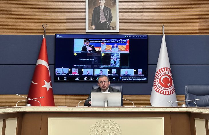 Görselde, TBMM’de bir toplantı salonu görülüyor. Ortada kürsüde oturan bir kişi bilgisayar başından çevrim içi toplantıyı yönetiyor. Arka planda büyük ekranda çok sayıda katılımcının yer aldığı video konferans görüntüsü bulunuyor; salonun iki yanında Türk bayrağı ve TBMM bayrağı yer alıyor.