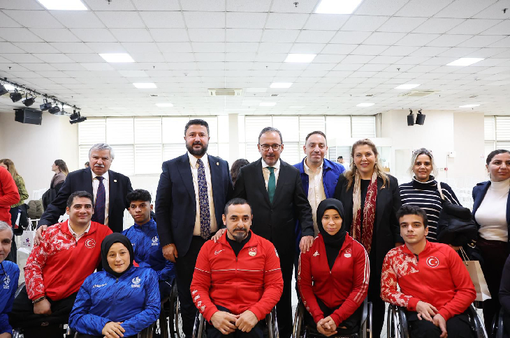 Geniş bir salonda, aralarında takım eşofmanları giyen tekerlekli sandalyeli sporcuların da bulunduğu engelli bireyler ve resmi kıyafetli yetkililer yan yana poz veriyor; kalabalık, düzenli ve resmi bir toplu fotoğraf.