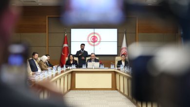 TBMM’deki bir toplantı salonu görülüyor. Uzun masanın etrafında milletvekilleri ve katılımcılar otururken, ortada konuşma yapan bir kişi var. Arkada Türk bayrağı ve TBMM amblemi yer alıyor; kadraj, resmî ve karar odaklı bir toplantı anını yansıtıyor.