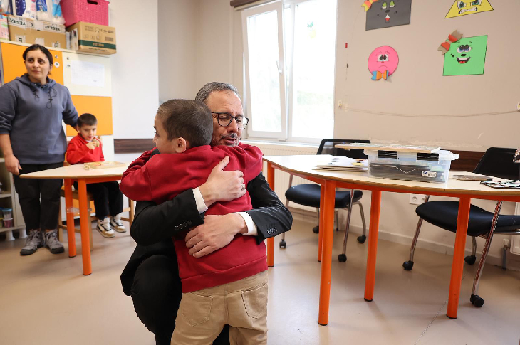 Sınıf ortamında, takım elbiseli bir yetkili küçük bir çocuğu kucaklayarak sarılıyor; arka planda öğretmenler, çocuklar ve renkli duvar süsleri görülüyor.