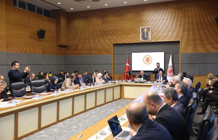 Türkiye Büyük Millet Meclisi’nde, uzun bir toplantı masası etrafında çok sayıda katılımcının yer aldığı resmi bir komisyon toplantısı görülüyor. Salonda Türk bayrakları, kürsü ve arka planda TBMM amblemi bulunurken, katılımcılar not alıyor ve konuşmacıyı dikkatle dinliyor.
