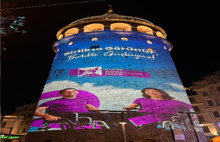 Görselde, Galata Kulesi’nin üzerine yansıtılmış büyük bir farkındalık görseli görülüyor. Işıklı projeksiyon üzerinde mor tonlarında tasarlanmış iki kişi –biri tekerlekli sandalyede– yer alıyor. Üst tarafta “Birlikte Görünür, Birlikte Güçlüyüz!” mesajı öne çıkıyor. Alt kısımda ilgili derneğin logosu bulunuyor. Gece çekilmiş renkli ve etkileyici bir projeksiyon çalışmasıdır.