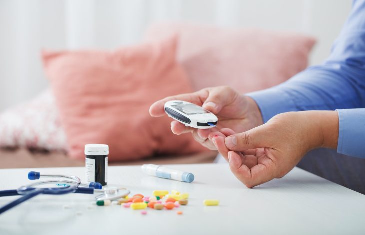 Fotoğrafta bir kişi ev ortamında kan şekeri ölçümü yapıyor. Elindeki glukometreyle parmağından ölçüm alırken masanın üzerinde çeşitli ilaçlar, haplar, bir enjeksiyon kalemi ve stetoskop bulunuyor. Arka planda yumuşak tonlarda minderler ve bulanık bir oturma odası detayı görülüyor.