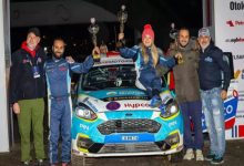 Görselde, bir ralli aracının önünde yan yana duran altı kişilik bir ekip görülüyor. Ortadaki üç kişi, araç üzerine çıkarak kazandıkları kupaları havaya kaldırmış. Hepsi yarış tulumları veya spor kıyafetler içinde, mutlu ve gururlu bir poz veriyor. Aracın üzerindeki yarış logoları ve sponsor etiketleri dikkat çekiyor.