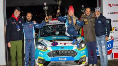 Görselde, bir ralli aracının önünde yan yana duran altı kişilik bir ekip görülüyor. Ortadaki üç kişi, araç üzerine çıkarak kazandıkları kupaları havaya kaldırmış. Hepsi yarış tulumları veya spor kıyafetler içinde, mutlu ve gururlu bir poz veriyor. Aracın üzerindeki yarış logoları ve sponsor etiketleri dikkat çekiyor.