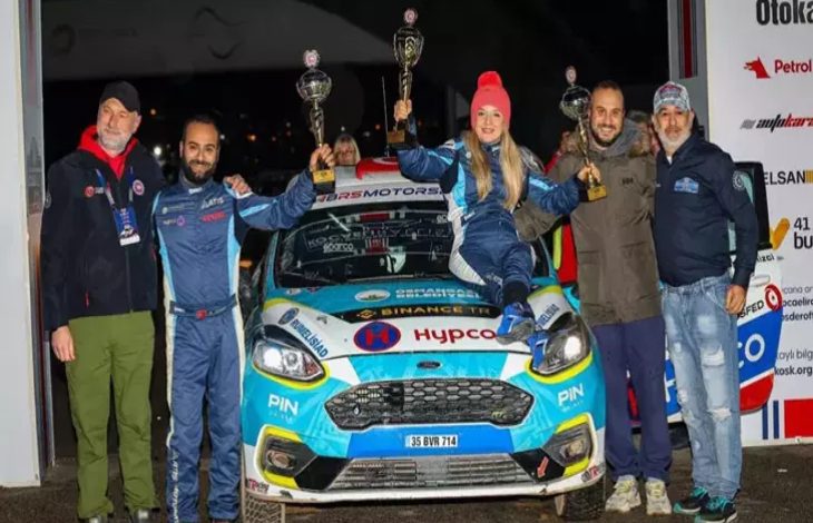 Görselde, bir ralli aracının önünde yan yana duran altı kişilik bir ekip görülüyor. Ortadaki üç kişi, araç üzerine çıkarak kazandıkları kupaları havaya kaldırmış. Hepsi yarış tulumları veya spor kıyafetler içinde, mutlu ve gururlu bir poz veriyor. Aracın üzerindeki yarış logoları ve sponsor etiketleri dikkat çekiyor.