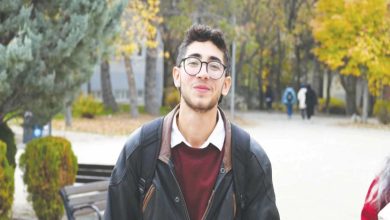 Görselde, açık havada bir kampüs yolunda yürüyen genç bir erkek görülüyor. Üzerinde koyu renk mont ve sırt çantası var, gözlük takıyor ve hafif gülümseyen bir ifadeye sahip. Arka planda sonbahar renklerine bürünmüş ağaçlar, banklar ve uzakta yürüyen insanlar yer alıyor.