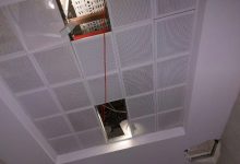 Fotoğrafta, tavanı oluşturan kare formlu, delikli panellerden biri yerinden çıkmış ve tavandaki kablolar ile yapı elemanları açık şekilde görülüyor; ayrıca bir aydınlatma armatürü de boşluktan aşağı doğru ışık veriyor.