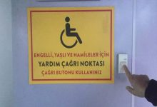 Görselde, mor renkte bir duvar üzerinde **engelli, yaşlı ve hamile bireyler için hazırlanmış bir yardım çağrı noktası** bulunuyor; sarı zeminli ve kırmızı çerçeveli uyarı tabelesinin yanında ise bir kişinin parmağı, **çağrı butonuna** basmak üzere uzanmış durumda.