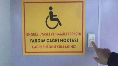 Görselde, mor renkte bir duvar üzerinde **engelli, yaşlı ve hamile bireyler için hazırlanmış bir yardım çağrı noktası** bulunuyor; sarı zeminli ve kırmızı çerçeveli uyarı tabelesinin yanında ise bir kişinin parmağı, **çağrı butonuna** basmak üzere uzanmış durumda.