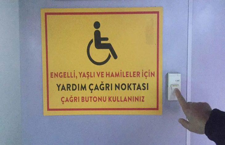 Görselde, mor renkte bir duvar üzerinde **engelli, yaşlı ve hamile bireyler için hazırlanmış bir yardım çağrı noktası** bulunuyor; sarı zeminli ve kırmızı çerçeveli uyarı tabelesinin yanında ise bir kişinin parmağı, **çağrı butonuna** basmak üzere uzanmış durumda.