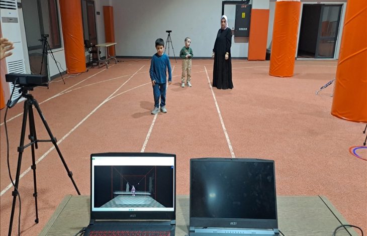 Geniş bir iç mekânda atletizm pistlerini andıran çizgiler var. Ortada, farklı yaşlarda iki çocuk ve arkalarında siyah uzun elbiseli bir yetişkin kadın ayakta duruyor. Ön planda bir masa üzerinde iki dizüstü bilgisayar bulunuyor. Bilgisayarlardan birinin ekranında, hareket yakalama/simülasyon benzeri bir görüntüyü gösteren bir sahne açık.