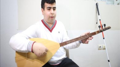 Bir sınıf ortamında oturan görme engelli genç, bağlamayı dizlerine yaslayıp dikkatle çalıyor; yanında beyaz bastonu dururken sahne sade ama umut dolu bir çalışma anını yansıtıyor.