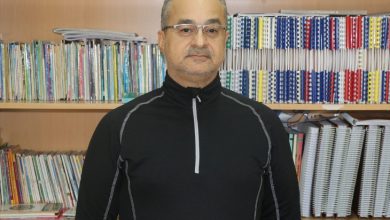 Raflarla dolu bir kütüphane önünde, gözlüklü bir adam kameraya dönük şekilde duruyor. Arkasında renk renk kitaplar ve dosyalar sıralanmış. Duruşu sakin, ortam akademik ve ciddi.