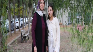 Ağaçların altında, park benzeri bir alanda yan yana duran iki kişi görülüyor. Biri başörtülü yetişkin bir kadın, diğeri açık renk kıyafetli genç bir kız; kameraya dönük, sakin ve vakur bir şekilde poz veriyorlar. Arka planda ağaçlar, bank ve park halindeki araçlar yer alıyor.