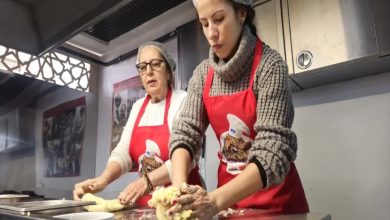 Endüstriyel bir mutfakta iki kadın, başlarında bone ve üzerlerinde kırmızı önlüklerle hamur yoğuruyor. Paslanmaz tezgâhın üzerinde un ve hamur parçaları var.