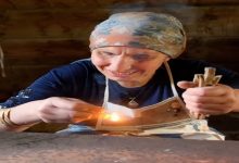 Ahşap bir masada, başörtülü yaşlı bir kadın gülümseyerek küçük bir ateşi üflüyor. Elinde çubuklar ve önünde yanan bir parça; yüzündeki çizgiler hem emek hem hikâye anlatıyor.