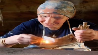 Ahşap bir masada, başörtülü yaşlı bir kadın gülümseyerek küçük bir ateşi üflüyor. Elinde çubuklar ve önünde yanan bir parça; yüzündeki çizgiler hem emek hem hikâye anlatıyor.