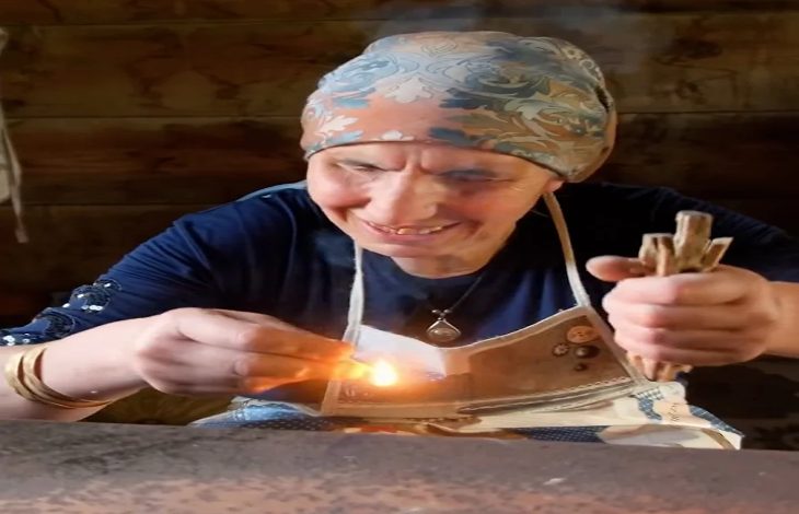 Ahşap bir masada, başörtülü yaşlı bir kadın gülümseyerek küçük bir ateşi üflüyor. Elinde çubuklar ve önünde yanan bir parça; yüzündeki çizgiler hem emek hem hikâye anlatıyor.