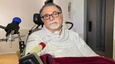 ALS hastalığıyla mücadele eden emekli göz doktoru Alper Kaya, tekerlekli sandalyede oturuyor, boynunda tıbbi bir solunum aparatı bulunuyor ve üzerinde açık renkli bir kazak var.