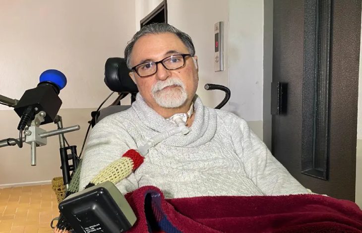 ALS hastalığıyla mücadele eden emekli göz doktoru Alper Kaya, tekerlekli sandalyede oturuyor, boynunda tıbbi bir solunum aparatı bulunuyor ve üzerinde açık renkli bir kazak var.