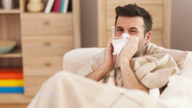 Ev ortamında koltukta oturan bir erkek, battaniyeye sarılmış halde mendille burnunu siliyor; grip ya da soğuk algınlığına bağlı halsiz bir an yansıtılıyor.