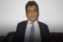 Açık renk bir arka plan önünde oturan, takım elbise ve kravatlı orta yaşlı bir erkek; kameraya sakin ve ciddi bir ifadeyle bakıyor.