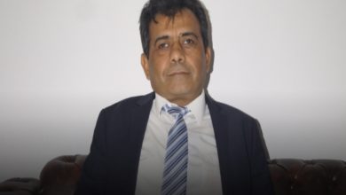 Açık renk bir arka plan önünde oturan, takım elbise ve kravatlı orta yaşlı bir erkek; kameraya sakin ve ciddi bir ifadeyle bakıyor.