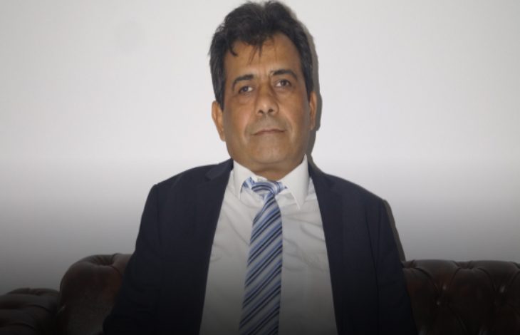 Açık renk bir arka plan önünde oturan, takım elbise ve kravatlı orta yaşlı bir erkek; kameraya sakin ve ciddi bir ifadeyle bakıyor.