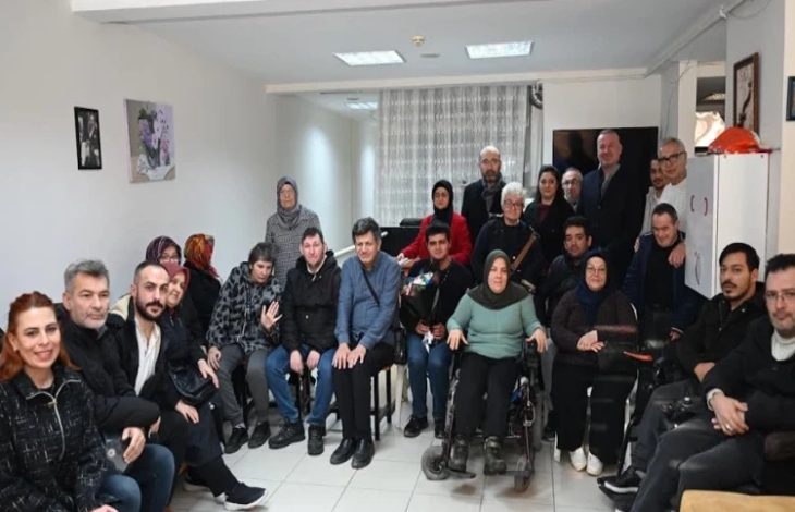 Kalabalık bir odada farklı yaşlardan kadın ve erkekler bir araya gelmiş, çoğu sandalyede ya da tekerlekli sandalyede oturuyor. Grup kameraya dönük, samimi ama ciddi bir toplu fotoğraf pozu veriyor. İç mekân sade; duvarda resimler, arkada perde ve ışıklar var. Ortam, dayanışma ve birlikte olma hissi taşıyor.