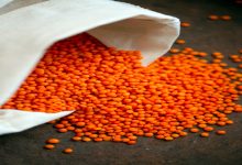 Beyaz bir bez torbadan dökülen parlak turuncu mercimek taneleri, koyu renk bir zemin üzerinde dağılmış halde görülüyor.