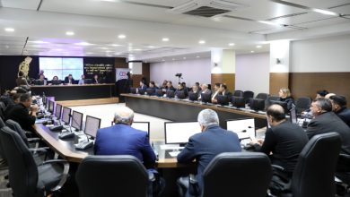 Modern bir toplantı salonunda, U şeklinde dizilmiş masalar etrafında bilgisayarlar önünde oturan Belediye Meclis üyeleri toplantı yapıyor.