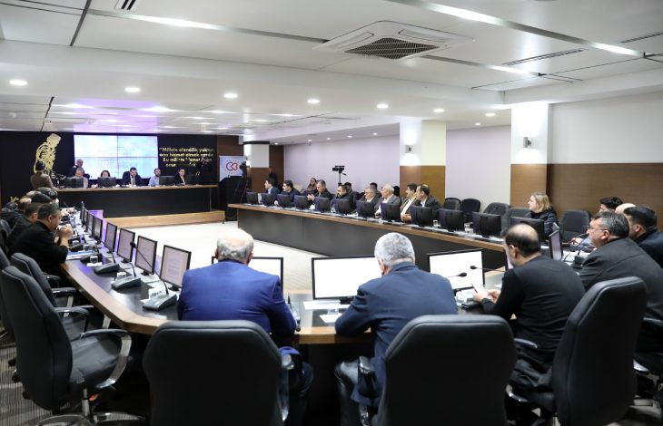 Modern bir toplantı salonunda, U şeklinde dizilmiş masalar etrafında bilgisayarlar önünde oturan Belediye Meclis üyeleri toplantı yapıyor.