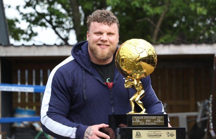 Tom Stoltman, açık havada spor kıyafetleriyle dururken elinde altın renkli, küre taşıyan bir heykelcikten oluşan büyük bir kupa tutuyor.