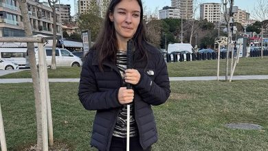 Açık bir park alanında genç bir kadın, elinde beyaz bastonuyla kameraya dönük duruyor. Siyah montlu, hafif gülümsüyor. Arkada çimenler, ağaçlar ve şehir binaları uzanıyor. Gün ışığı net ve sakin.
