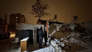 Gece vakti, karla kaplı bir avluda hasar görmüş bir ev görülüyor. Duvarları kısmen yıkılmış, çatısı çökmüş. Kapı önünde iki görevli ayakta duruyor; içeride yaşanan felaketin sessiz tanıkları gibi.