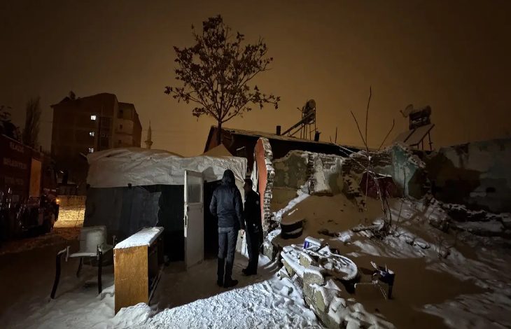 Gece vakti, karla kaplı bir avluda hasar görmüş bir ev görülüyor. Duvarları kısmen yıkılmış, çatısı çökmüş. Kapı önünde iki görevli ayakta duruyor; içeride yaşanan felaketin sessiz tanıkları gibi.