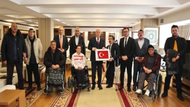 Geniş bir salonda kalabalık bir grup poz veriyor. Ortada takım elbiseli bir yetkili, elinde Türk bayrağı bulunan çerçeveyi tutuyor. Ön sırada tekerlekli sandalyedeki bireyler yer alıyor; arkada ise ayakta duran diğer katılımcılar.