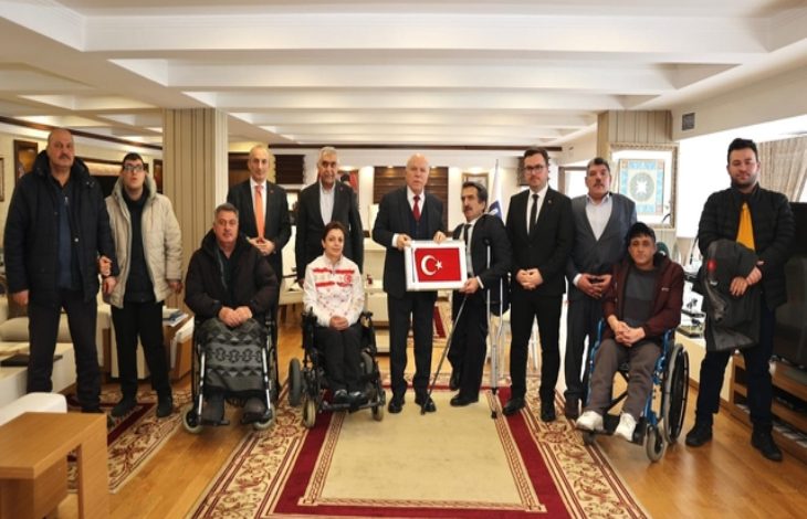 Geniş bir salonda kalabalık bir grup poz veriyor. Ortada takım elbiseli bir yetkili, elinde Türk bayrağı bulunan çerçeveyi tutuyor. Ön sırada tekerlekli sandalyedeki bireyler yer alıyor; arkada ise ayakta duran diğer katılımcılar.