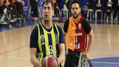 Bir spor salonunda tekerlekli sandalyede basketbol oynayan iki sporcu görülüyor. Önde topu elinde tutan sporcu şuta hazırlanırken, arkasındaki rakip oyuncu savunmada; maçın ciddiyeti ve rekabet duygusu öne çıkıyor.