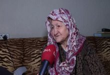 Evinde röportaj veren yaşlı bir kadın, başörtüsüyle koltukta otururken DHA mikrofonuna konuşuyor; yüzündeki ifade yaşadığı zorluğu ve yardım beklentisini yansıtıyor.