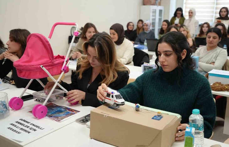 Sınıf ortamında, sıralara dizilmiş öğrenciler dikkatle kendi geliştirdikleri maket projelerle ilgileniyor. Ön planda iki genç kadın; biri pembe oyuncak bebek arabasıyla, diğeri karton bir platform üstünde küçük bir ambulans modeliyle çalışıyor. Masada su şişeleri, notlar ve proje kartları var.